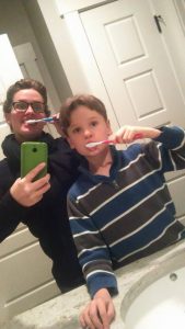 An adult and a child stand in front of a bathroom mirror brushing their teeth. The adult holds up a green phone to take a selfie. Both are smiling and appear to be enjoying the moment together.