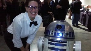 A smiling person wearing glasses, a white shirt, and a black bow tie stands next to an R2-D2 robot replica at an indoor event with other people in the background.