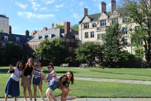 Five smiling young women pose playfully on a sunlit lawn with historic campus buildings and leafy trees in the background, under a blue sky with wispy clouds.