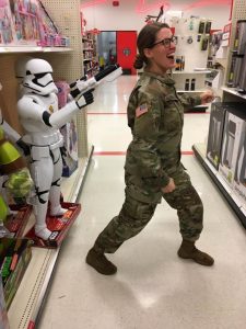 A woman in military uniform playfully pretends to be held up by a life-size Stormtrooper figure pointing a toy blaster at her in a store aisle. She is smiling and striking a dramatic pose.