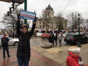 A woman in a winter coat holds a Refugees Welcome sign at a street protest, with a large crowd and courthouse building in the background on a cloudy day.