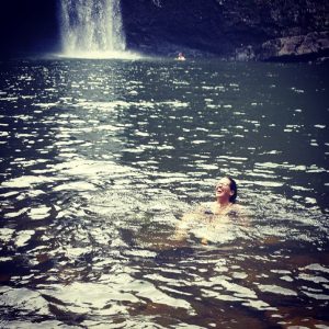 A person joyfully swimming in a lake near a waterfall, with lush rocks and water splashing around. The waterfall cascades in the background, adding to the serene, natural setting.