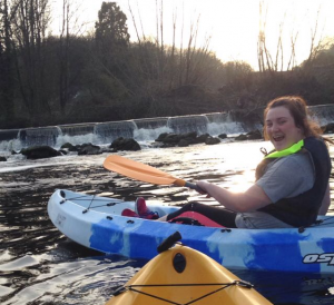 A smiling young woman in a blue kayak paddles on a river near a small waterfall, wearing a life vest. The water is calm and trees line the riverbank in the background at sunset.