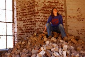 A person in a blue shirt and jeans sits atop a large pile of bricks inside a weathered, brick-walled room with a tall window on the left side. They look upward with a thoughtful expression.