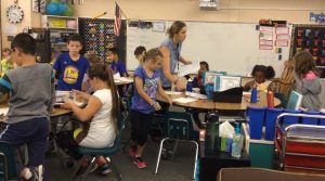 A classroom scene with several elementary students working at tables and a teacher walking around assisting them. The classroom is colorful, with various supplies, posters, and an American flag on the wall.