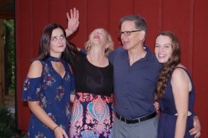 Four people stand together in front of a red wall. One woman is laughing with her head back and arm raised, while the others smile; a young woman on the left rolls her eyes playfully. They appear to be a family.