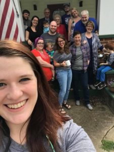A group of people, including adults and children, smile for a selfie in front of a house with an American flag and a porch. A woman in the foreground holds the camera, and everyone appears happy and relaxed.