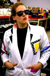 A woman wearing black sunglasses and a bold, colorful, patterned blazer poses confidently with her hands in her pockets inside a store, standing in front of racks of clothes and other merchandise.