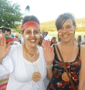 Two women smiling and waving at an outdoor event, with face paint and feather headbands. Both wear name tags and summer clothes. There are other people in the background, also dressed festively.