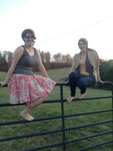 Two smiling women sit barefoot on a metal fence in a grassy field, with trees and hills in the background. One wears a floral skirt and tank top; the other wears pants, a yellow shirt, and a light jacket.