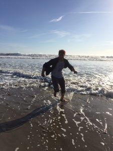A person runs out of the shallow waves onto a sandy beach, casting a long shadow behind them. The sky is clear and blue with a few clouds, and the ocean waves are gently crashing in the background.