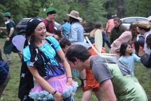 A group of people dressed in colorful costumes, some with fairy wings, laugh and dance outdoors on grass, surrounded by trees and parked cars. The atmosphere is lively and festive.