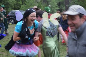 A group of people in colorful costumes, including fairy wings and tutus, laugh and dance outdoors on grass with trees and parked cars in the background. The mood appears joyful and festive.