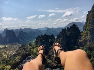 A person wearing sandals sits with legs outstretched, overlooking a scenic mountain landscape with rocky peaks, green trees, and a blue sky with scattered clouds.