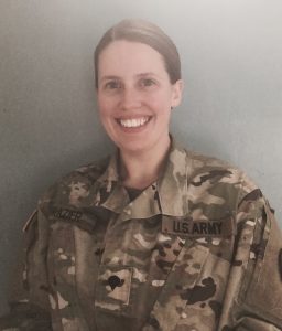 A woman in a U.S. Army uniform with a camouflage pattern smiles at the camera. She has light brown hair pulled back and stands against a plain light blue background. The name tag reads FRAZIER.