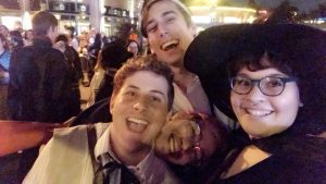 Four friends smile and pose for a selfie at a lively outdoor event at night. One person wears a wide-brimmed witch hat and glasses, while the others are dressed casually and appear joyful in the crowd.
