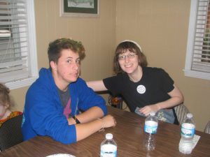 Two people sit at a wooden table with bottled water and a paper plate. One person wears a blue hoodie, the other a black shirt and white headband, smiling and reaching toward the first person. Theres a window in the background.