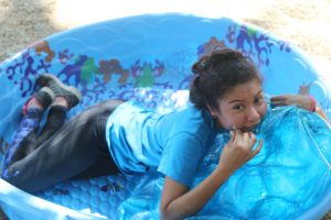 A young person in a blue shirt and black pants lies in a plastic kiddie pool, playfully biting an inflated blue pool float. The scene appears fun and relaxed, and the pool is decorated with colorful patterns.