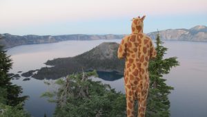 A person wearing a giraffe costume stands on a forested overlook, gazing out at a large, calm lake surrounded by cliffs and distant mountains under a clear sky.