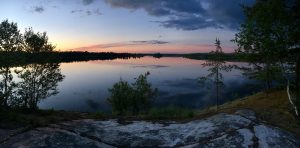 A tranquil lake at sunset with calm water reflecting the sky and clouds, surrounded by trees and rocky shoreline, under a colorful sky fading from blue to orange and pink.