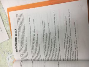 A close-up of a printed page titled FACILITATION SKILLS lists soft and hard skills, behaviors, and examples of good and bad leadership. The page rests on a folder, with a partially visible map underneath.