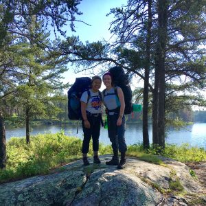Two people wearing backpacks and hiking gear stand together on a rocky area near a lake, surrounded by tall trees and greenery under a clear blue sky.