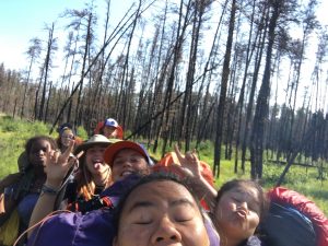 A group of smiling and laughing young people with backpacks take a selfie while hiking through a forest with tall, thin trees and green grass. Some make playful hand signs and funny faces.