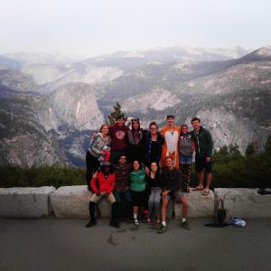 A group of twelve people pose and smile in front of a scenic mountain landscape with tree-covered slopes and distant peaks under a hazy sky. They appear relaxed and happy, some kneeling and others standing together.