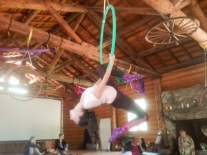 A person hangs upside down from a green aerial hoop in a rustic wooden room, wearing colorful socks. Several people sit or kneel on the wooden floor, watching. Sunlight streams in through the windows.