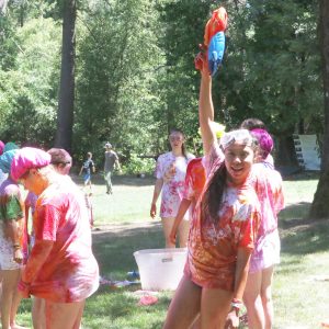A group of people in colorful, paint-splattered clothes stand outdoors on grass. One person in front, smiling, holds a red and blue water gun high, wearing goggles. Others stand nearby, also covered in paint. Trees are in the background.