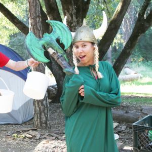 A person in a green outfit and Viking helmet with blond braids poses outdoors, holding a large green foam axe. Trees and a tent are in the background; another person’s arm holding two white buckets is visible on the left.