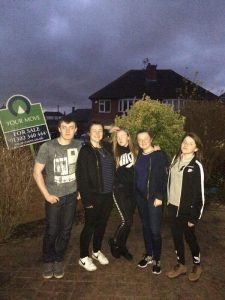 Five teenagers stand together outside in front of a house at dusk. A “For Sale” sign is visible on the left. The group poses casually, some with hands in pockets, with bushes and a dark, cloudy sky in the background.