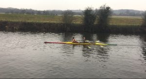 Two people are rowing a yellow double scull boat on a calm river, with grassy banks and trees in the background on a cloudy day.