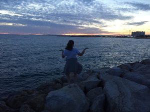 A person stands on rocks by the sea at sunset, facing the water with arms bent and raised. The sky is partly cloudy with orange and blue hues, and city lights are visible in the distance on the right.