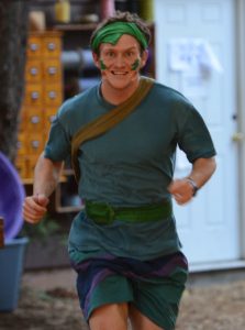 A person dressed in a green costume with face paint and a headband is running outdoors, smiling energetically. There is a white door and various objects blurred in the background.