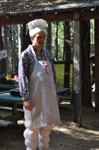 A person wearing a chef hat, apron, and padded pants stands outdoors in a wooded area, with sunlight filtering through trees and a rustic shelter in the background.