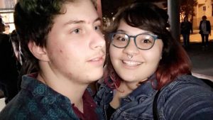 Two people posing for a selfie at night; one has short hair and is looking away, while the other, wearing glasses and a septum ring, is smiling at the camera. There are blurred people and lights in the background.