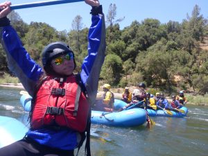 A person wearing a helmet, sunglasses, and a red life jacket hangs from a blue bar over a river. In the background, groups of people in helmets paddle blue rafts surrounded by trees under a clear sky.