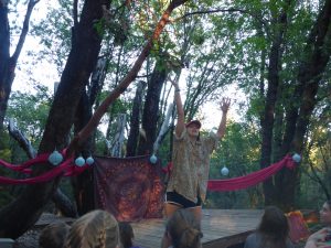 A person stands on an outdoor wooden stage, raising their arms. They wear a patterned shirt and shorts. Behind them, red fabric and lanterns are draped among trees. Several people watch from the audience.