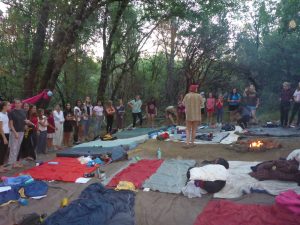A group of people, mostly young women, stand in a circle in a forest clearing at dusk. Sleeping bags and blankets are spread on the ground, and a small campfire burns nearby. Trees surround the campsite.