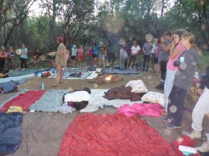 A group of people stands in a circle around sleeping bags and blankets laid on the ground in a wooded outdoor area, with a small campfire in the center and trees in the background.