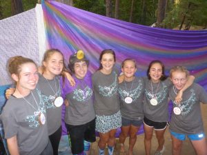 Seven smiling teenagers wearing matching gray shirts and handmade necklaces stand arm-in-arm in front of a colorful purple and blue fabric backdrop outdoors, surrounded by trees.