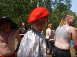 A person wearing a red beret, round glasses, and a shiny silver cape stands outdoors, surrounded by other casually dressed people. Trees and sunlight are visible in the background.