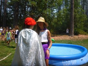 Two people stand near a blue inflatable pool outdoors; one wears a red beret and silver cape, the other wears a straw hat and purple shorts. Other children and trees are visible in the background.