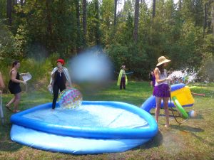 Four women enjoy a backyard scene with an inflatable pool and water toys. One woman stands in the pool with a beach ball, another sprays water, and two others hold pool noodles. Trees and greenery surround them.