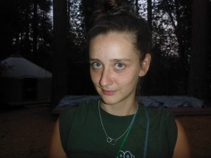 A young woman with light skin, green eyes, and brown hair in a bun stands outdoors at dusk, wearing a dark green shirt and a silver necklace. Tall trees and a tent are visible in the background.