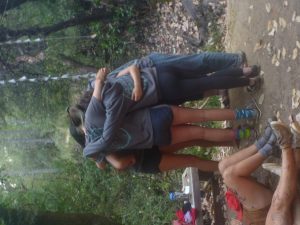 Five people in casual clothes stand in a group hug on a dirt clearing in the woods, while another person with writing on their legs sits nearby. Leaves scatter the ground and trees surround the scene.