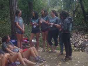 A group of young women stand and laugh together outdoors in a wooded area, holding round objects, while others sit or lie on the ground, surrounded by trees and fallen leaves.