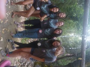 Six young women wearing matching dark t-shirts and round badges stand together in a forest, posing playfully and smiling for the camera. Leaves and trees fill the background.