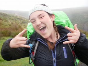 A smiling young woman in outdoor gear with a green backpack makes rock and roll hand signs. She wears a gray knit headband and stands in a foggy, grassy landscape.